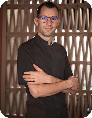 Chef en tenue noire, bras croisés, devant une paroi texturée, illustrant son expertise et son professionnalisme.